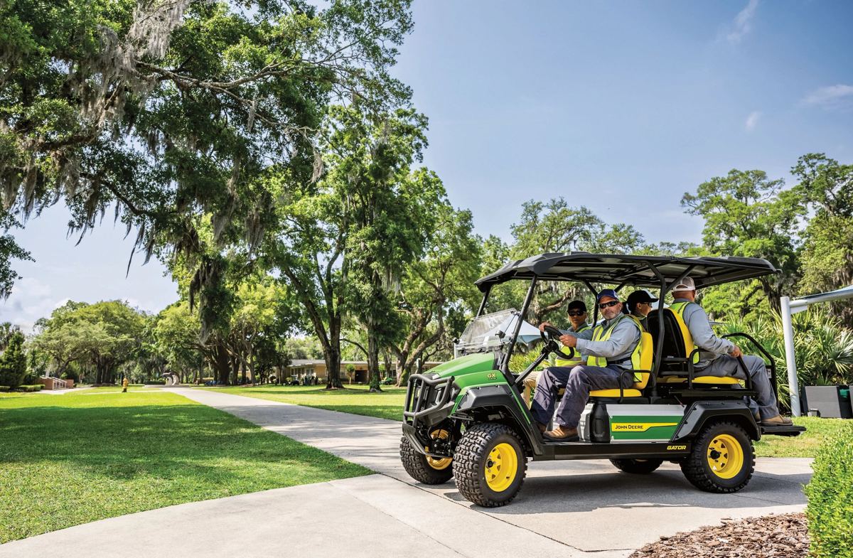 Four men in a green John Deere Gator utility vehicle on a paved path lined with trees
