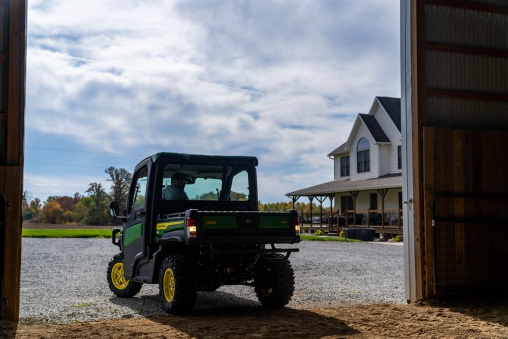 A John Deere Gator utility vehicle pulling out of a barn toward a farmhouse