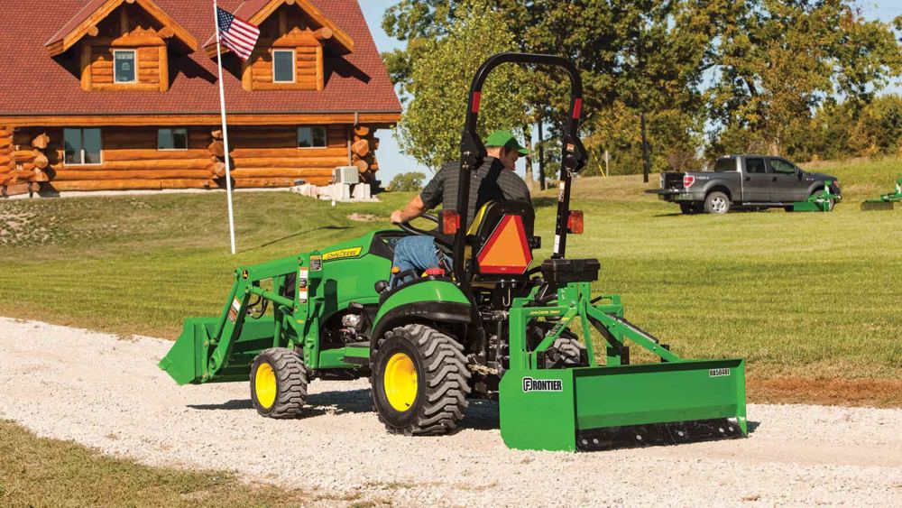 A John Deere compact tractor grading a gravel driveway with a box blade