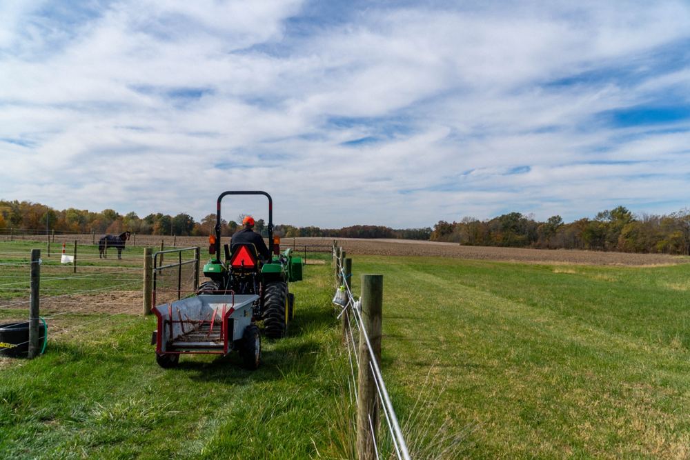 A John Deere compact tractor pulling an implement along a horse pasture fence line