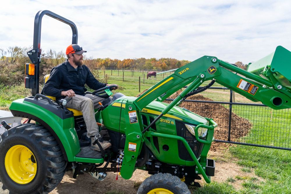 A John Deere compact tractor with a loader moving material beside a horse pasture