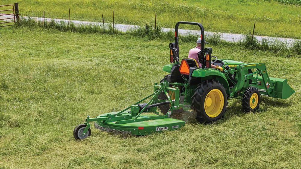 A John Deere compact tractor mowing tall pasture grass with a rotary cutter