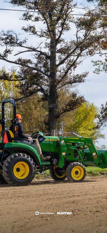 John Deere compact utility tractor with a front loader pulling an arena groomer past autumn trees, phone wallpaper preview