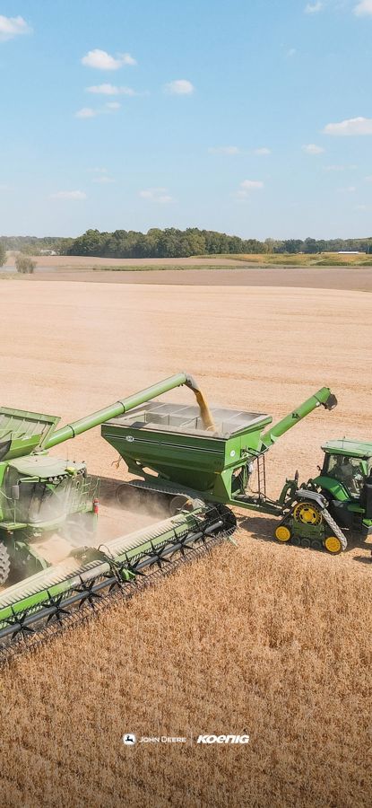 An aerial view shows a combine harvester unloading grain into a grain cart pulled by a tractor in a field under a blue sky., phone wallpaper preview